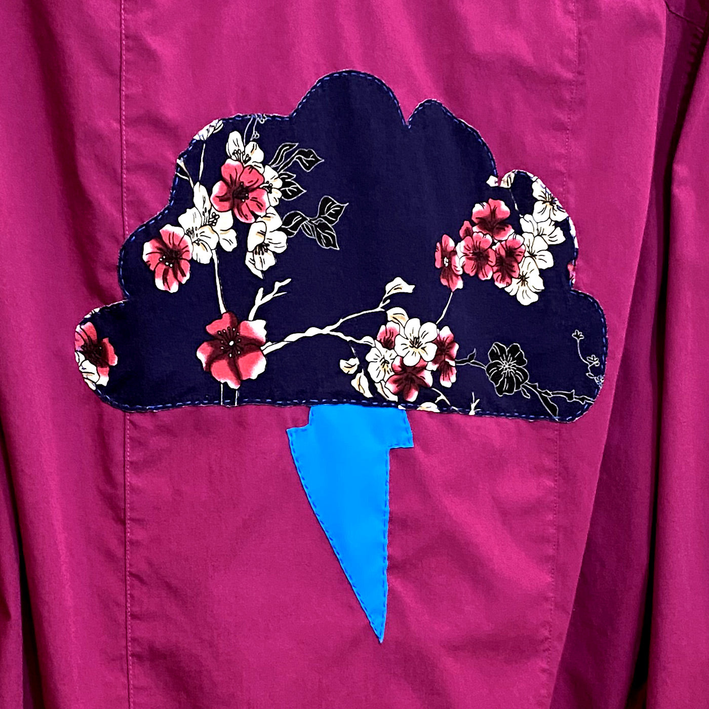 Close up of hand stitched navy cloud with red and cream flowers and leaves pattern with a light blue lightening bolt below the cloud