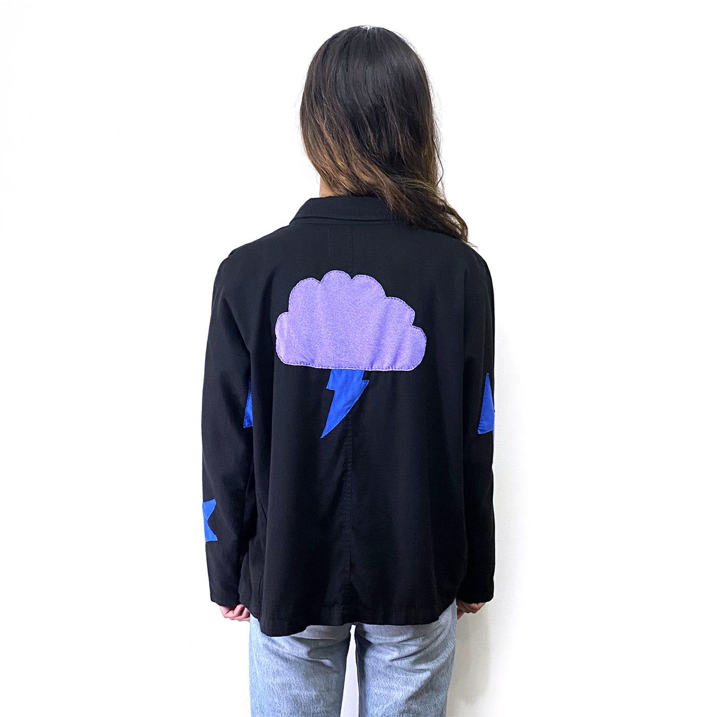 Back of young person with long brown hair wearing a short black jacket with a light purple cloud sewn on the centre of the back and a few blue lightening bolts included in the hand stitched design